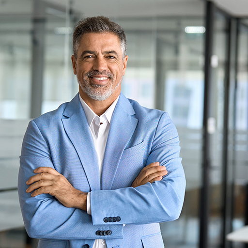 A grey-bearded man in a light blue blazer standing with his arms folded