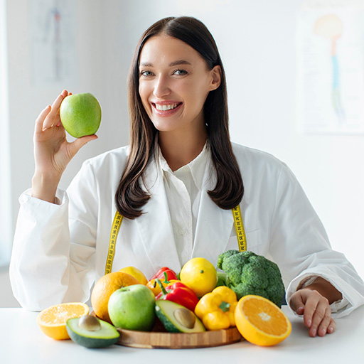A dietitian in a white coat holding an apple beside a bowl of fresh fruit and vegetables