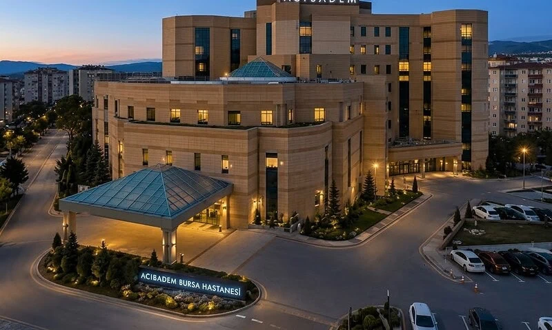 Exterior view of Acibadem Bursa Hospital at dusk with cityscape in background.