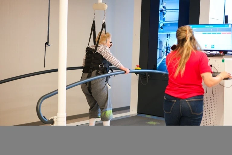 A patient walking on a treadmill in a body-weight support harness while a therapist runs the session from the console beside them