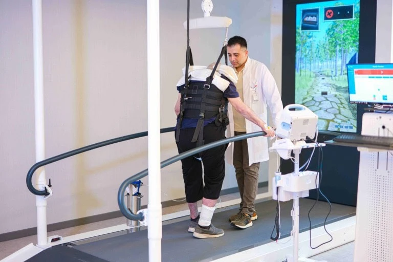 A patient walking on the Lokomat treadmill in a body-weight support harness while a therapist adjusts the control unit, with a virtual walking path on the screen ahead