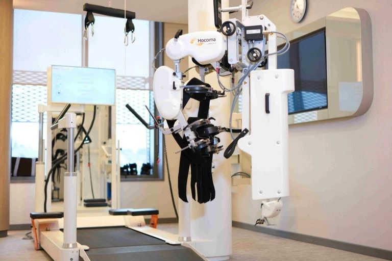 Lokomat robotic gait trainer with its robotic leg orthoses and overhead body-weight support harness, positioned over a treadmill in the rehabilitation gym